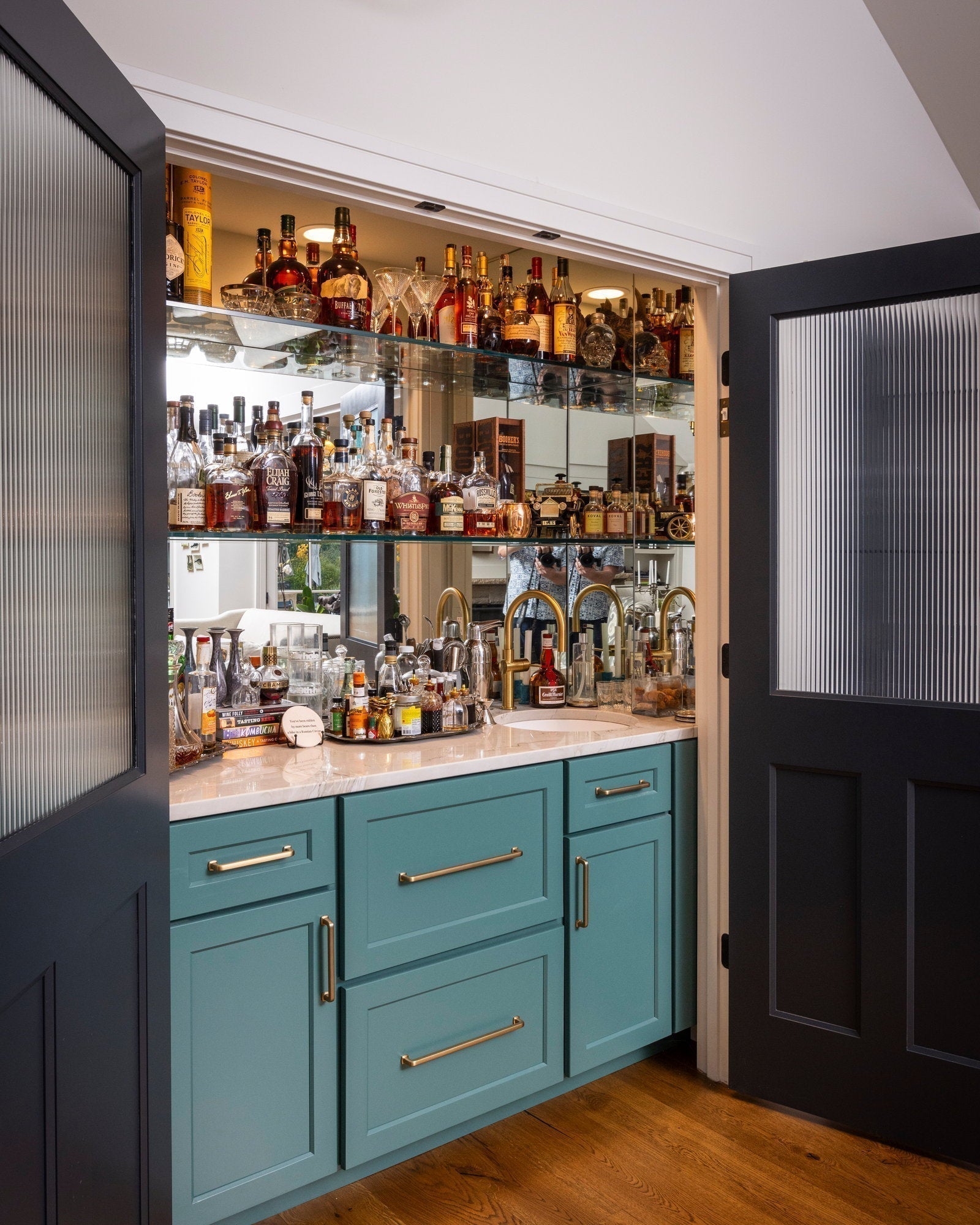 Navy reeded-glass doors open on a mirrored bar with lit shelves of bourbon, brass faucet, stone counter
