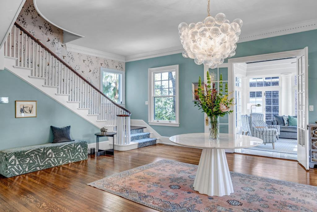 Teal entry hall with a white pedestal table, upholstered bench, glass chandelier and curved stair