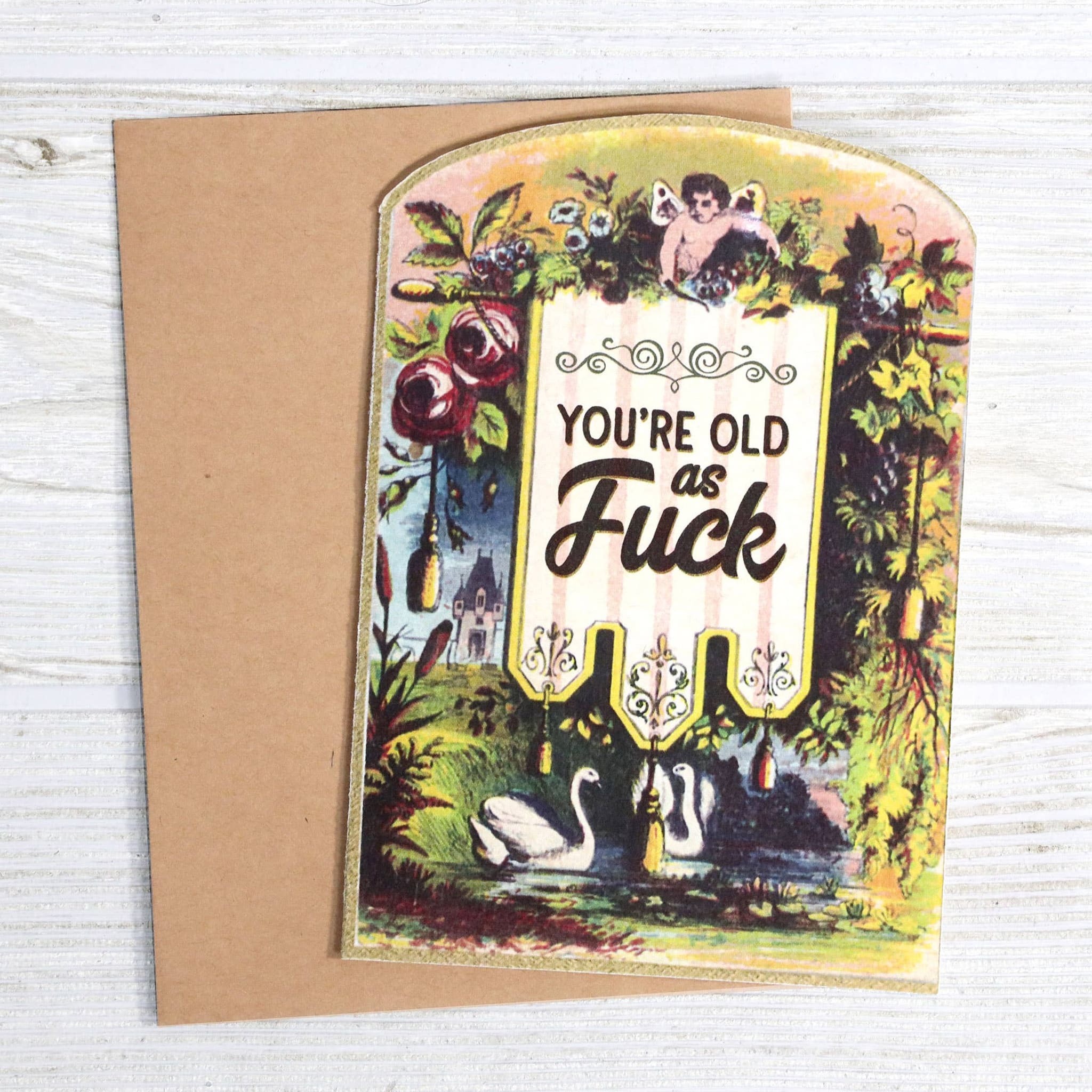 Greeting card with vintage botanical illustration, swans, cherub, and humorous birthday text, with kraft envelope on white wood surface