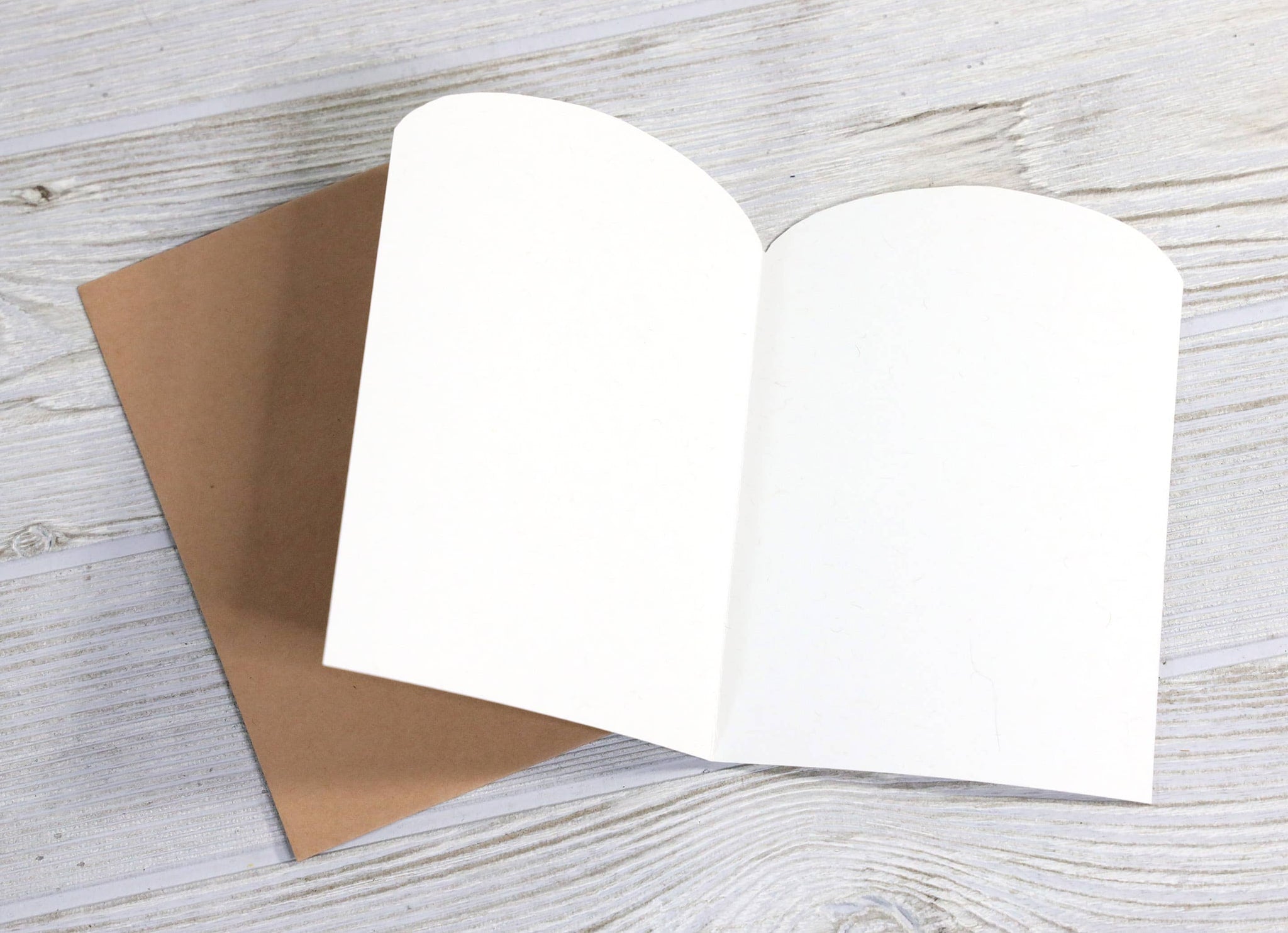 Greeting card open flat showing blank white interior pages with kraft brown cover on whitewashed wood surface