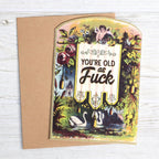 Greeting card with vintage botanical illustration, swans, cherub, and humorous birthday text, with kraft envelope on white wood surface