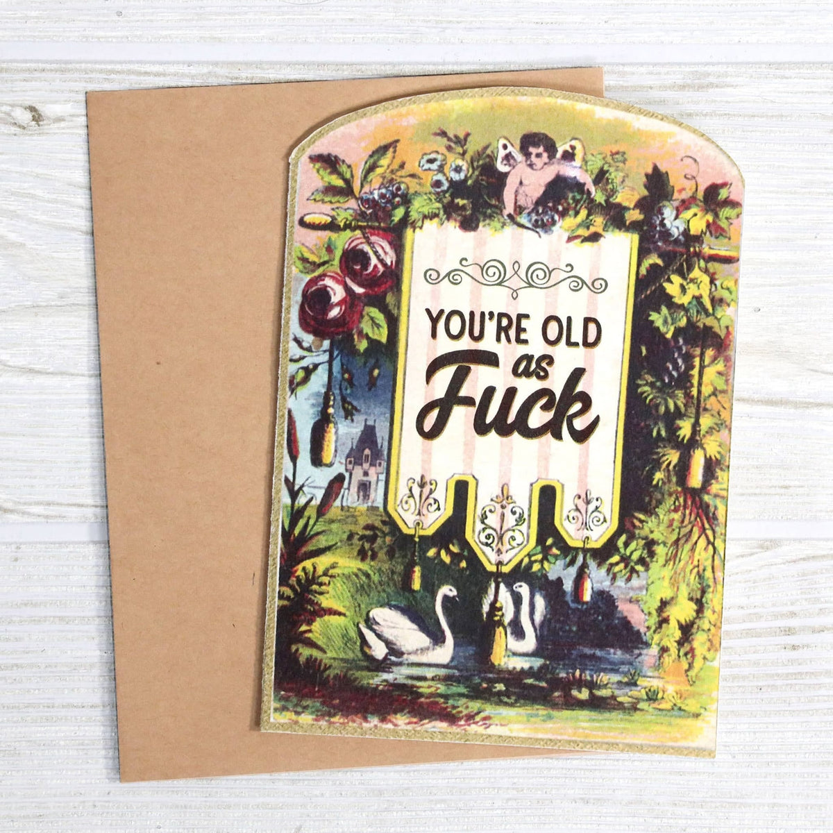 Greeting card with vintage botanical illustration, swans, cherub, and humorous birthday text, with kraft envelope on white wood surface