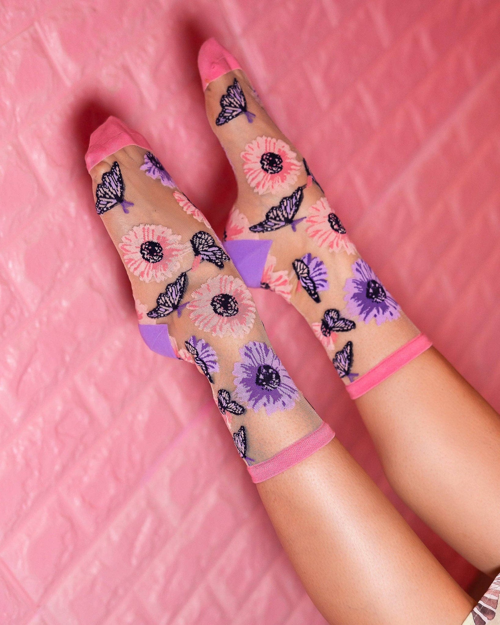 Sheer crew socks with pink and purple floral and butterfly print, worn on raised feet against pink background