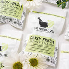 Shower steamer pouches, white with green label, surrounded by daisy flowers and greenery