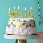 Gold balloon-letter "BLOW ME" birthday candles lit on white floral decorated cake
