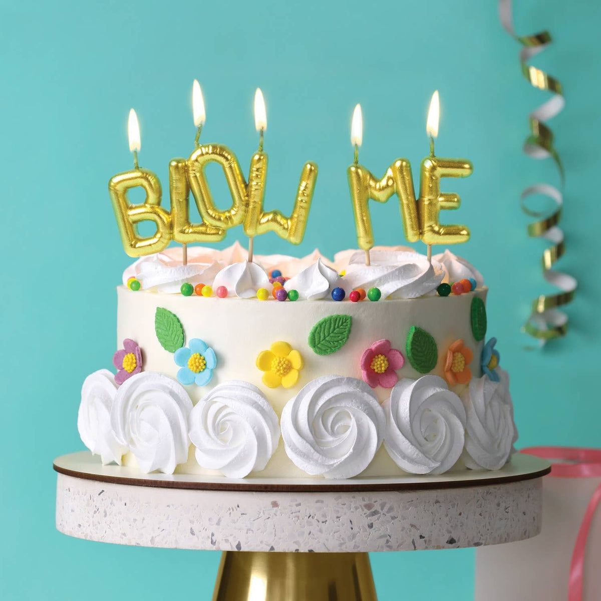 Gold balloon-letter "BLOW ME" birthday candles lit on white floral decorated cake