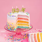Gold "OLD AF" letter balloon birthday candles lit on rainbow layer cake with sprinkles