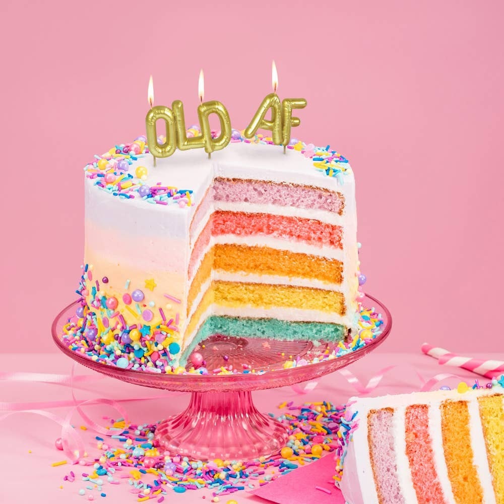 Gold "OLD AF" letter balloon birthday candles lit on rainbow layer cake with sprinkles