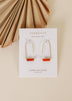 Earrings, gold arch drop style with orange carnelian agate bar, displayed on white card, top-down view