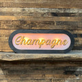 Lighted Champagne sign, dark charcoal frame, glowing pink backlit panel with gold script lettering, displayed on rustic wood shelf