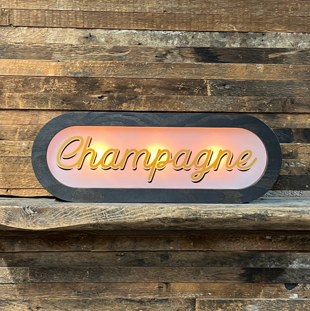 Lighted Champagne sign, dark charcoal frame, glowing pink backlit panel with gold script lettering, displayed on rustic wood shelf