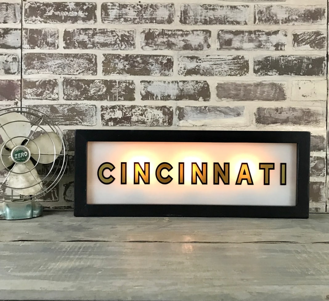 Lighted Cincinnati sign, black rectangular frame, illuminated gold letters, on wooden surface