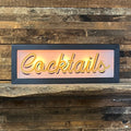 Lighted Cocktails sign, gold script lettering, dark gray frame, warm pink glow, mounted on rustic wood wall