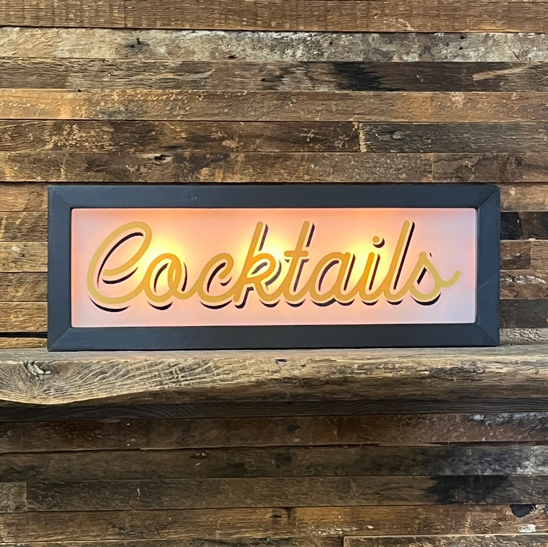 Lighted Cocktails sign, gold script lettering, dark gray frame, warm pink glow, mounted on rustic wood wall