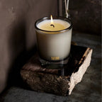 Candle in frosted glass vessel with lit flame, cream wax, gold Tatine label, on rustic wood slab