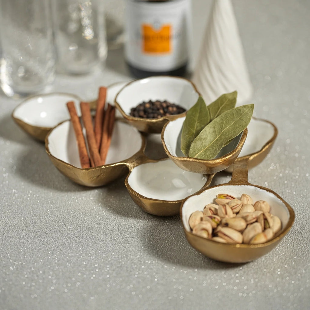 Gold and white ceramic cluster bowls holding cinnamon sticks, peppercorns, bay leaves, and pistachios on a glittery grey surface